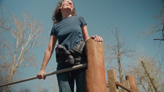 Close up of woman performing outdoor exercise on wooden equipment holding onto metal bars and wood posts. Woman wears casual clothing with item around her waist as woman moves
