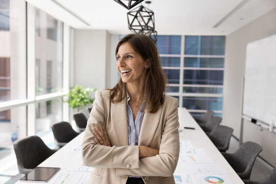 Smiling adult Latina female corporate executive standing with arms crossed in modern conference room looking forward career growth or project success showing leadership mindset professional motivation
