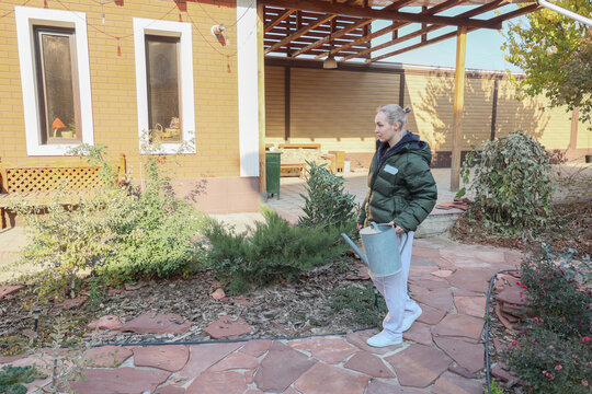 Middle Aged Woman Stands Beside Yellow Home In Landscaped Backyard, Holding Blue Watering Can Near Shrubs And Stone Path, With Pergola Overhead, Windows, Patio Area And Soft Autumn