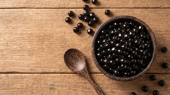 Discover top-view flatlay of freshly prepared chewy black tapioca pearls in a wooden bowl and spoon on a rustic wooden table, perfect for bubble tea.