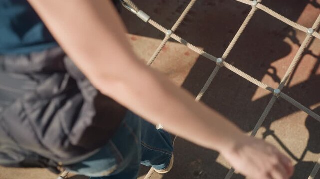 Close up of person legs and feet as they carefully step across outdoor rope bridge balancing on thick ropes with metal connectors. person wears blue jeans and dark sneakers while navigating