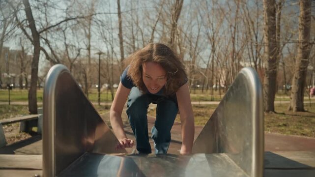 Close up of young woman running towards camera in outdoor park then climbing and interacting with playground equipment. Woman wears casual clothing including jeans and t shirt throughout