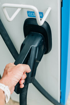 Closeup of an electric vehicle charging connector plugged into a public charging station with a hand holding the cable