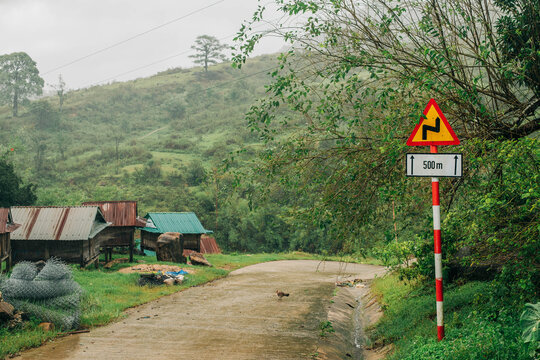 Winding mountain road with a warning sign and rural huts during heavy rain