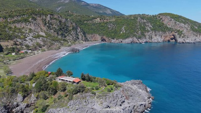 Drone footage capturing a unique house perched atop a rocky cliff, overlooking the golden sandy beach and crystal-clear waters of Limnionas Beach on Evia Island, Greece.