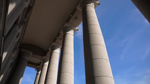 Sweeping wide angle shot captures grand neoclassical columns and ornate capitals under clear blue sky, showcasing dramatic architecture in natural daylight with strong upward perspective.