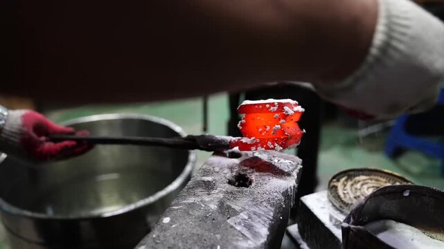 Glowing damascus billet coated with borax flux on an anvil next to a steel quenching bowl in a knife workshop