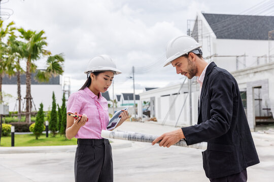 Female architect using a digital tablet while a male engineer reviews a rolled blueprint at a residential construction site. Professional teamwork