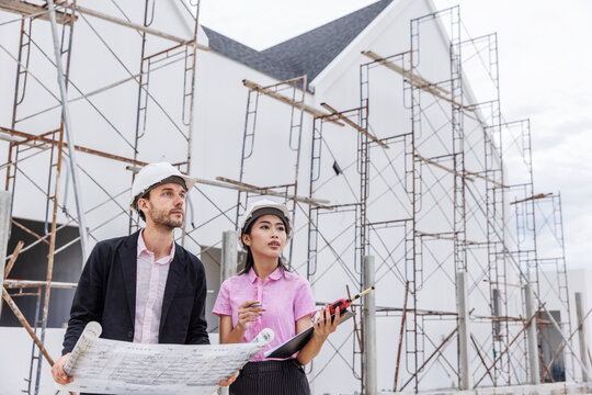 Male engineer and female architect wearing safety helmets while reviewing an architectural blueprint beside scaffolding at a building construction site.