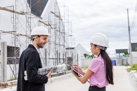 Male engineer and female architect wearing safety helmets while reviewing an architectural blueprint beside scaffolding at a building construction site.
