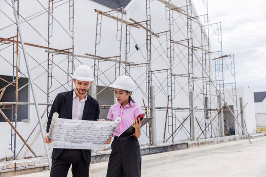 Male engineer and female architect wearing safety helmets while reviewing an architectural blueprint beside scaffolding at a building construction site.