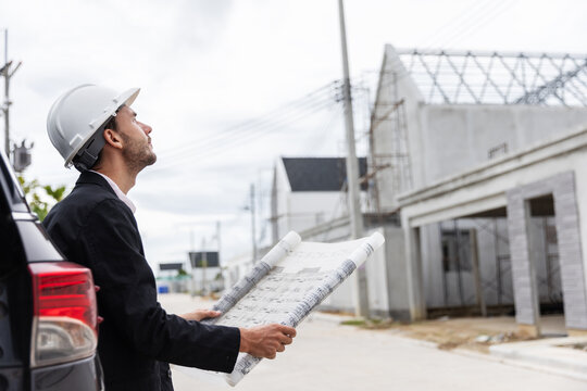 Professional male engineer wearing a safety helmet and holding an architectural blueprint while inspecting the progress of a residential construction project. 