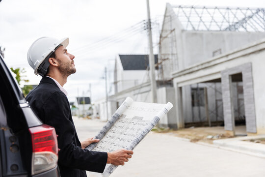 Professional male engineer wearing a safety helmet and holding an architectural blueprint while inspecting the progress of a residential construction project. 