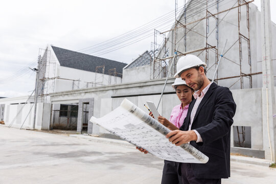 Male engineer and female architect wearing safety helmets while reviewing a large construction blueprint at a residential development site. Professional teamwork, architectural planning