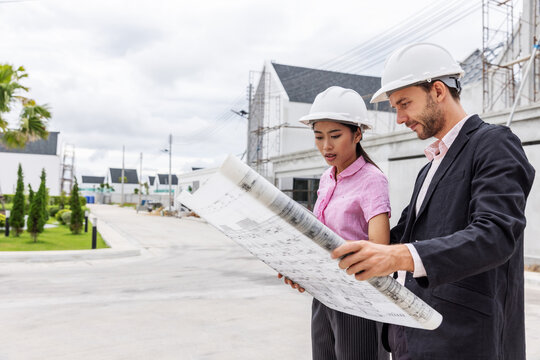 Male engineer and female architect wearing safety helmets while reviewing a large construction blueprint at a residential development site. Professional teamwork, architectural planning
