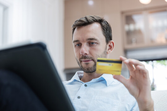 Young man holding a credit card while using a laptop to make an online purchase from home. E-commerce checkout, secure digital payment, internet banking, cashless transaction and consumer technology .