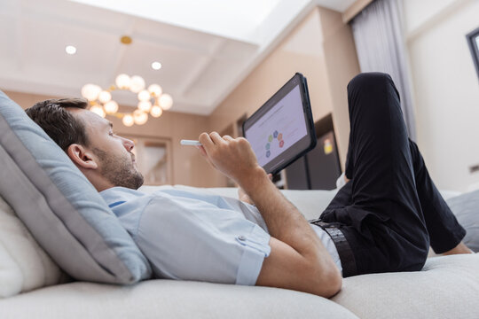 Young businessman relaxing on a sofa while reviewing financial charts and business analytics on a digital tablet in a modern living room. 