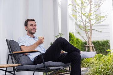 Fototapeta premium Smiling young man enjoying a cup of coffee while relaxing on a comfortable chair in a modern outdoor patio and green garden.