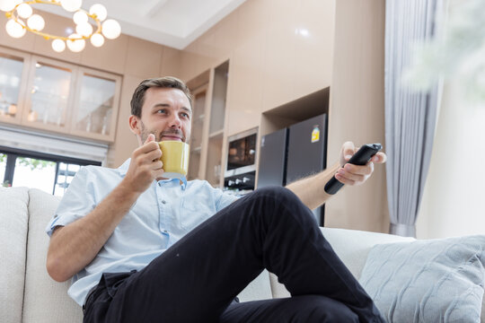 Smiling young man holding a remote control and enjoying a hot drink while relaxing on a comfortable sofa in a modern living room.