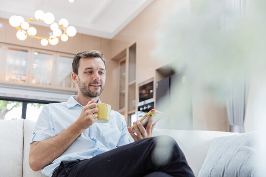 Smiling young man checking his smartphone while enjoying a cup of coffee on a comfortable sofa in a modern living room. Mobile communication, social media, online lifestyle, leisure and relaxation.