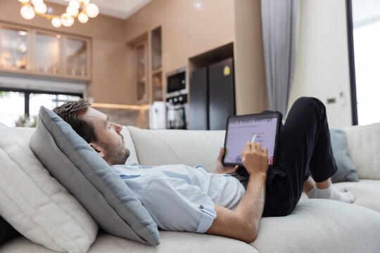 Young businessman relaxing on a sofa while reviewing financial charts and business analytics on a digital tablet in a modern living room. 