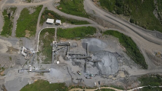 Aerial view of a stone quarry featuring conveyor belts, piles of crushed rock, and heavy machinery in the rural landscape of Rumpin, West Java, Indonesia.