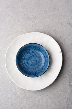 Top view of minimalist white ceramic plates and a deep blue bowl arranged on a gray textured stone countertop with a white linen napkin. Neutral table setting for dining, kitchen, home decor concept