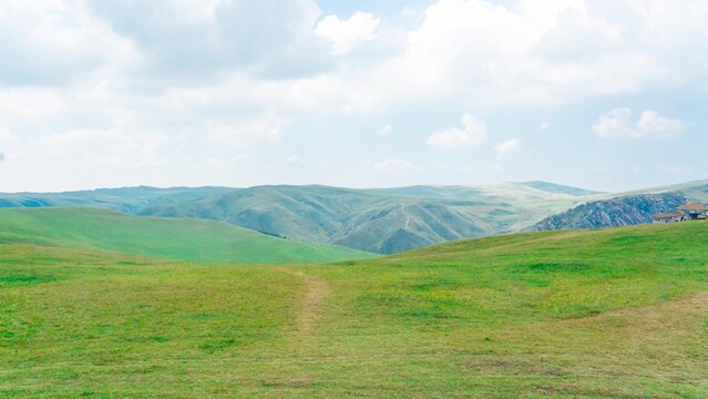 Scenic green grassland with a dirt path leading to rolling hills and blue sky with clouds