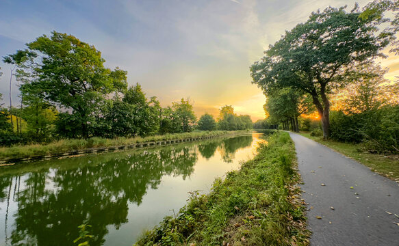 golden sunrise over mirrorlike canal surface with towpath and trees, warm light bathes water, reeds and soft mist create tranquil