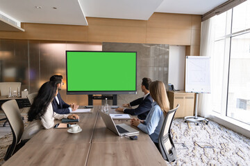 Obraz premium Group of businesspeople sit at conference table, watching presentation, engaging in video call, looking at digital screen displaying green mockup during corporate business meeting in office boardroom