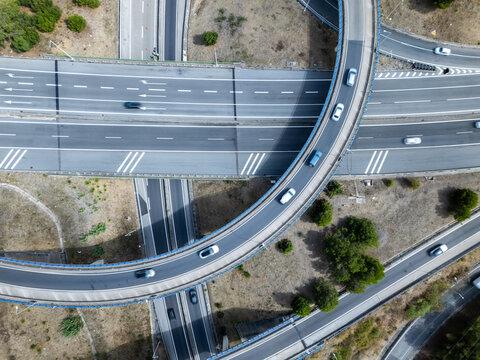 Aerial view of the A5 highway interchange with vehicles driving along curved ramps and multi-level roads surrounded by dry vegetation in Cascais, Lisboa, Portugal.