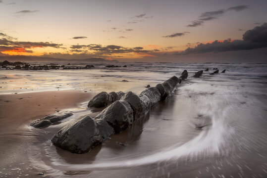Flysch rock formations sticking out from the wet sand on the beach of Barrika at sunset with low shutter speed water motion, Basque Country, Spain