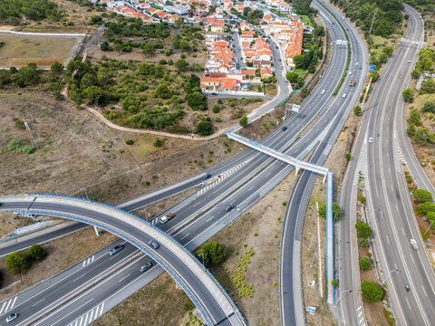 Aerial view of the A5 highway interchange with vehicles on the asphalt, a pedestrian bridge, and surrounding dry vegetation and suburban housing in Cascais, Lisboa, Portugal.