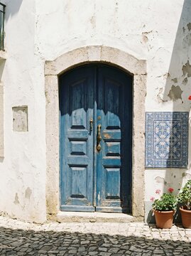 Narrow cobblestone street with whitewashed buildings, blue wooden door and pink flowers, blue tiles near sea view at Amalfi coast.