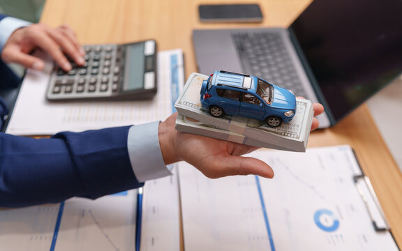 Businessman holds a model car on cash above financial documents near a calculator and laptop, representing car loans, vehicle financing, credit approval, payments and purchase planning.