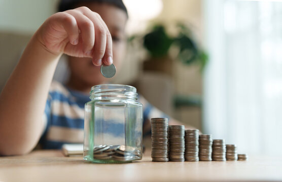 Asian Child drops a coin into a glass savings jar beside growing stacks of coins at home, representing saving habits, financial education, money management, future planning and savings growth.