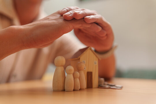 Hands shelter wooden family figures and a house with coins on a table, symbolizing family protection, home insurance, financial security, property coverage, risk management and future planning.