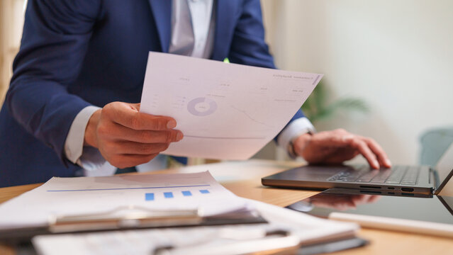 Businessman reviews a financial report while working on a laptop at an office desk, representing financial analysis, audit, compliance, document inspection, reporting and data accuracy.
