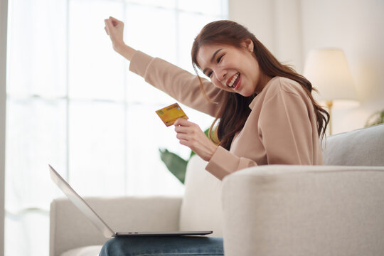 Happy Asian woman celebrates a successful online payment while holding a credit card beside a laptop at home, representing e-commerce shopping, digital banking, cashless purchase and checkout success.