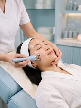 An Asian woman relaxes as a specialist uses a microcurrent facial massage device on her cheek, a skincare and beauty treatment concept.