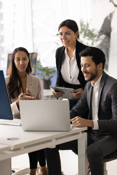 Smiling multiethnic professional business team collaborate around laptop, reviewing project data, discussing strategic decisions together, working on task during briefing in coworking office. Teamwork