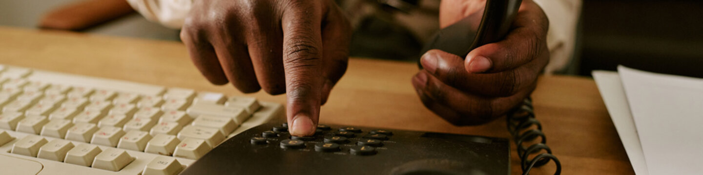 Header of hands of African American man dialing number on vintage landline telephone in retro office. Image represents corporate communication history and is suitable for business heritage marketing