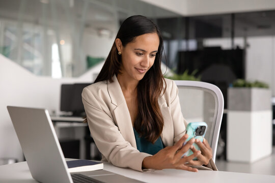 Adult New-Zealander businesswoman reviews messages, checking smartphone, making call seated at workplace office desk with laptop. Mobile application use, digital connectivity, business communication