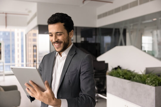Smiling Middle-eastern businessman working on digital tablet standing in corporate office, typing message, using business application, reviewing successful results, good news or positive report data