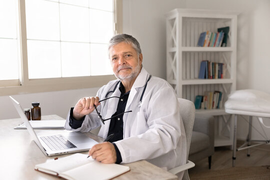Smiling senior male doctor sitting at workplace desk, looking at camera, representing professional healthcare services, workflow at hospital office, medical competence, and patient care, portrait