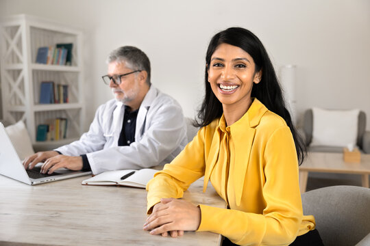 Portrait of South Asian woman patient sits at desk during visit at hospital while male doctor working on laptop, checking medical information, preparing treatment plan. Healthcare consultation concept
