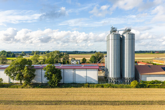 Aerial view of modern agricultural grain silos and farm buildings with solar panels on the roof, surrounded by cultivated fields under a blue sky in Gniew, Pomeranian Voivodeship, Poland.