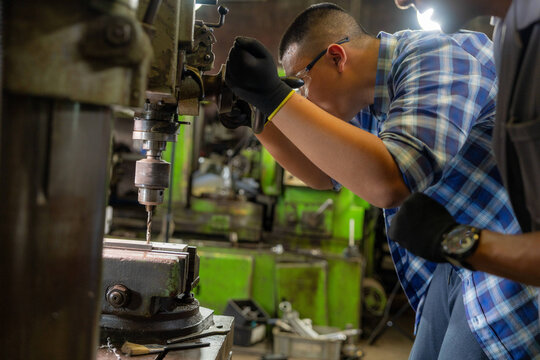 Teenage son of a factory owner inside the old family machine shop, son bends low over the drill table, right gloved hand deep under the spindle, next generation learning to inherit