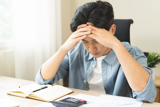 Debt and financial concept, stressed asian young man have a headache while male sitting calculate expenses, income, hand holding bills for prepare payment or pay bills on table at home.
