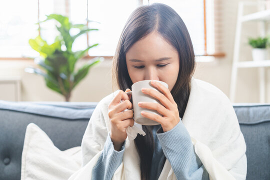 Sick concept, weakness asian young woman have a fever, flu and girl have a headache drinking a mug of warm water to keep the body warm while sitting rest on sofa at home. Health care on virus person.